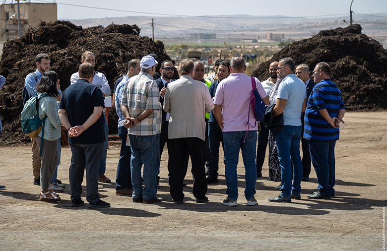 A group of people are standing in a circle listening to a man speaking. Behind them, you can see two mounds of compost.